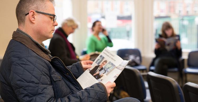 A man is sitting in a busy waiting room of a surgery and waiting to be seen. He is reading a service information booklet