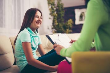 A girl is smiling and sitting on a sofa. She is in conversation with an adult who is holding a clipboard.