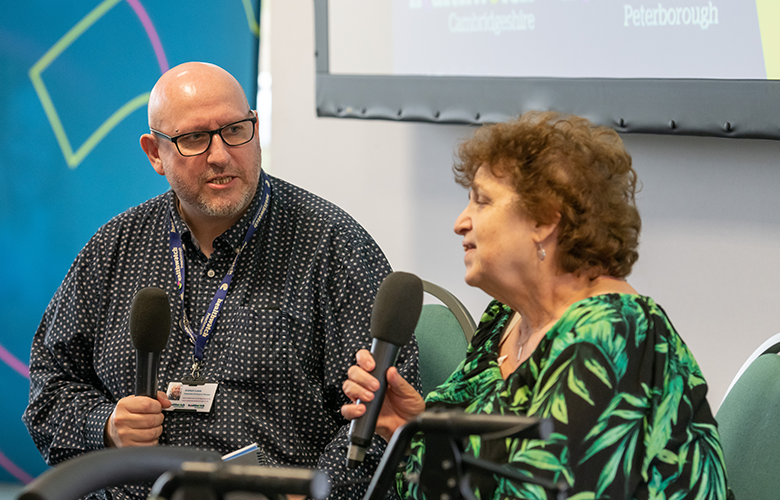 Woman and man sat on stage, Woman has microphone and is speaking 