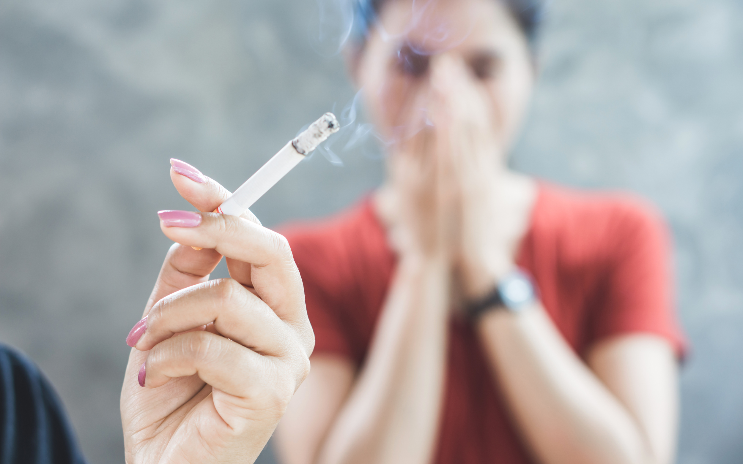 A woman holding a lit cigarette with someone in the background holding their hands over their face