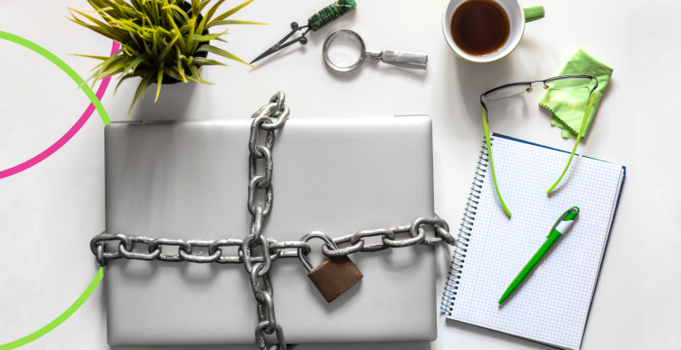 A photo of a laptop chained and locked with a padlock, next to a plant, cup of coffee and a notepad with pen.