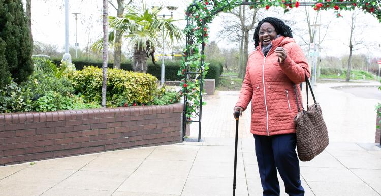 Woman with walking stick and carrying a bag. She is walking along a street.