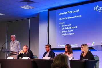 Our Question Time panel and host seated on a stage and listening to questions from the audience. Behind the panel is a large screen with their names on it.