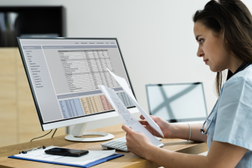 Photo of a woman sat at a computer and desk looking at data on sheets of paper