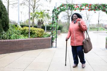 Woman with walking stick and carrying a bag. She is walking along a street.