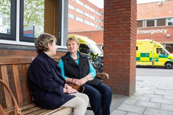 Two women sitting on a bench outside a hospital and talking to each other. There is an ambulance parked in the background.