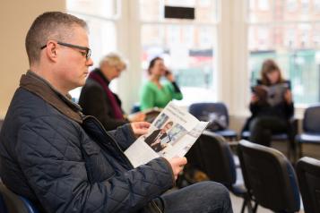 A man is sitting in a busy waiting room of a surgery and waiting to be seen. He is reading a service information booklet