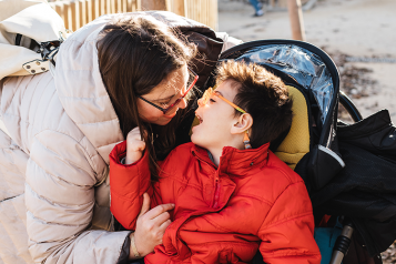 disabled boy in wheelchair outside wearing redcoat and woman leaning over to hug him