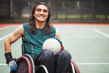 Smiling man in sportswear, sat in wheelchair on sports court holding ball.