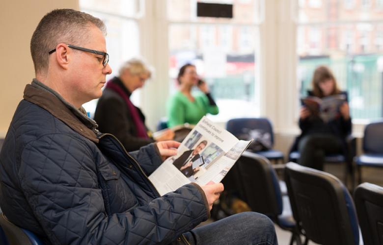 A man is sitting in a busy waiting room of a surgery and waiting to be seen. He is reading a service information booklet
